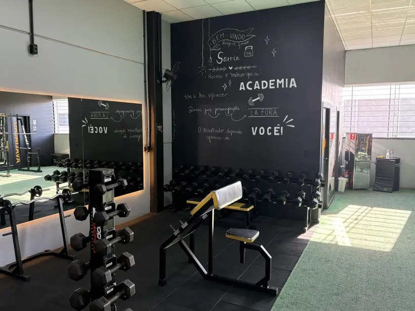 Rack de halteres e banco regulável ao lado do painel de lousa da academia.